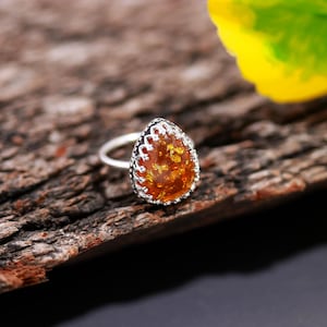 Könnte beinhalten: Silberring mit tropfenförmigem Bernstein. Der Bernstein hat Goldeinschlüsse und ist in einer dekorativen Silberfiligranfassung gefasst. Der Ring wird auf einem Holzstück präsentiert.