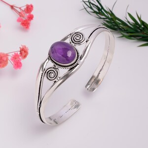 Könnte beinhalten: Ein silberfarbenes Armband mit einem zentralen, ovalen violetten Edelstein. Das Metallband ist auf beiden Seiten des Steins mit kunstvollen Drahtwirbeln verziert, was ein offenes Design ergibt, das zu den offenen Enden hin schmaler wird.