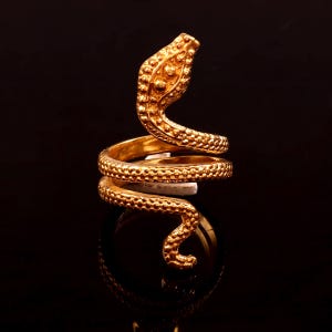 Può includere: Un anello color oro a forma di serpente, avvolto attorno al dito. La testa del serpente è sollevata, con squame dettagliate e una superficie strutturata. L'anello è posto su uno sfondo nero.