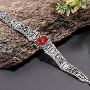 May include: A silver bracelet featuring an oval, reddish-orange stone. The bracelet has an ornate, filigree design with intricate patterns. The bracelet is displayed on a piece of wood.
