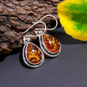 May include: A pair of silver teardrop earrings with amber-colored stones. The stones have a golden, textured appearance and are set in a silver frame with a twisted rope design. The earrings have a hook closure.