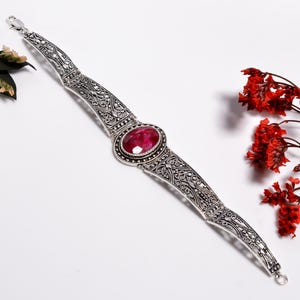 Pode incluir: Uma pulseira de metal prateado com uma pedra preciosa oval facetada de cor rosa profundo no centro. A pulseira é composta por elos de metal articulados de estilo filigrana com acabamento polido e um fecho de lagosta seguro.