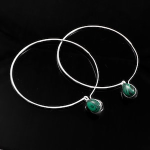 Könnte beinhalten: Zwei silberfarbene Halsketten mit kreisförmigem Design. Jede Halskette hat einen tropfenförmigen Anhänger aus grünem und schwarzem Stein. Der Schmuck wird vor einem schwarzen Hintergrund präsentiert, der den Kontrast von Farben und Texturen hervorhebt.