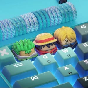 May include: A custom mechanical keyboard with light blue keycaps and a matching coiled cable. The keycaps feature character-themed artisan keycaps, including a green plant, a character with a straw hat, and a character with blonde hair.
