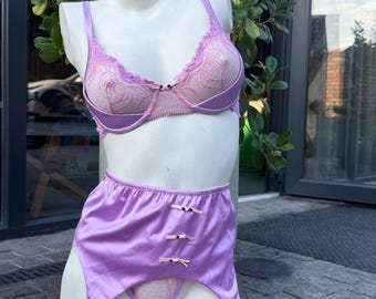 May include: A lavender lingerie set featuring a sheer lace underwire bra with scalloped edges, a high-waisted satin suspender belt with three small bows, and matching lace-trimmed hold-up stockings displayed on a white mannequin torso.