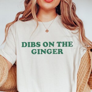 Może przedstawiać: Kremowy t-shirt z napisem "DIBS ON THE GINGER" w zielonych literach. Koszulka jest noszona z beżowym kardiganem i jasnoniebieskimi dżinsami. Ogólny styl jest swobodny i wygodny.