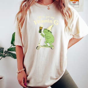 May include: A cream-coloured t-shirt featuring a cartoon frog on a scooter, holding a pint of beer. The text reads "Off to get my Vitamin G". The shirt is worn by a person. The frog wears a wizard hat.
