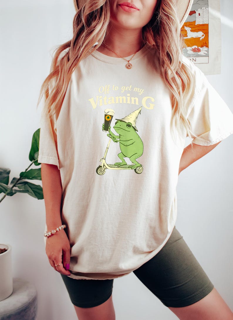 May include: A cream-coloured t-shirt featuring a cartoon frog on a scooter, holding a pint of beer. The text reads "Off to get my Vitamin G". The shirt is worn by a person. The frog wears a wizard hat.