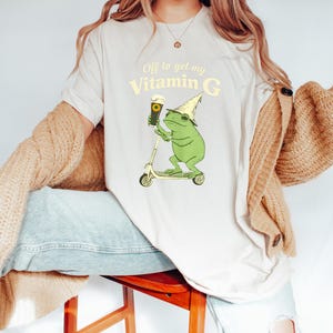May include: A cream-coloured t-shirt with a cartoon frog on a scooter, holding a beer and wearing a party hat. The text "Off to get my Vitamin G" is printed above the frog illustration in a light yellow colour.
