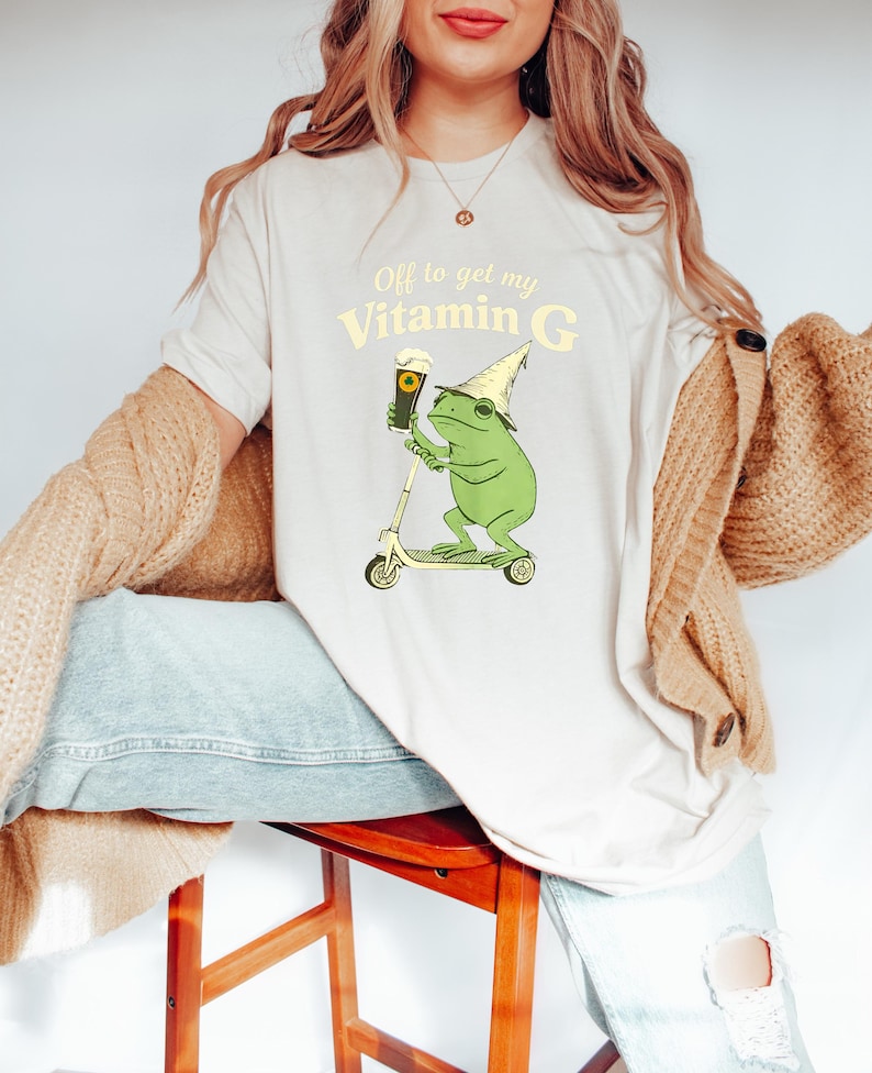May include: A cream-coloured t-shirt with a cartoon frog on a scooter, holding a beer and wearing a party hat. The text "Off to get my Vitamin G" is printed above the frog illustration in a light yellow colour.