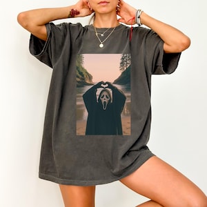 May include: Dark gray t-shirt featuring a graphic of a person in a Scream mask forming a heart shape with their hands. The image is set against a beach backdrop with a sunset. The shirt has short sleeves and a relaxed fit.