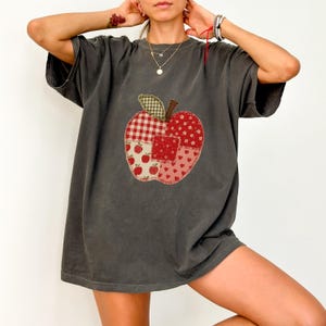 Op de afbeelding: Een antracietgrijs T-shirt met korte mouwen en een grote centrale appelafbeelding, samengesteld uit diverse rood-witte stoffen met patronen zoals vichy-ruitjes, bloemen en hartjes, met een groen geruit blaadje aan de bovenzijde.