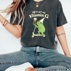May include: Dark grey t-shirt with the text "Off to get my Vitamin G." The shirt features a green frog riding a scooter and holding a pint of beer. The frog wears a wizard hat. The shirt is paired with blue jeans and white trainers.