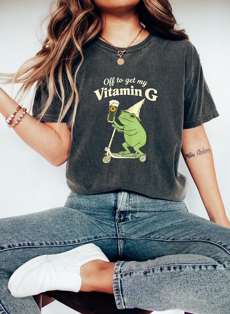 May include: Dark grey t-shirt with the text "Off to get my Vitamin G." The shirt features a green frog riding a scooter and holding a pint of beer. The frog wears a wizard hat. The shirt is paired with blue jeans and white trainers.