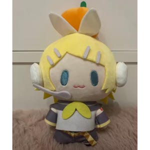 May include: A plush doll featuring a yellow-haired character with teal eyes, a small pink mouth, and a headset. The doll wears a gray and white outfit with a yellow bow and a small orange hat with a green stem. The doll is on a pink surface.