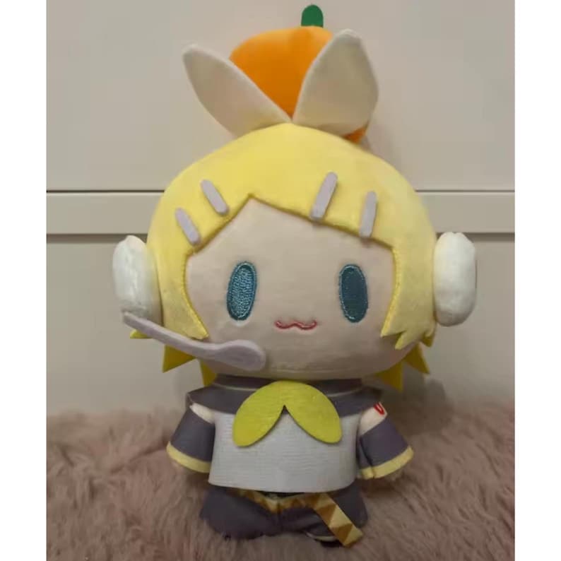 May include: A plush doll featuring a yellow-haired character with teal eyes, a small pink mouth, and a headset. The doll wears a gray and white outfit with a yellow bow and a small orange hat with a green stem. The doll is on a pink surface.