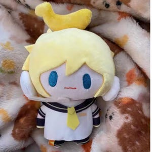 May include: A plush doll of a character with yellow hair and a yellow banana-shaped accessory. The doll has a white sailor uniform with a yellow tie and black accents. The doll has blue eyes and a small red mouth.