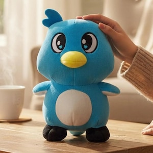 Pode incluir: Um brinquedo de pelúcia de pássaro azul com bico amarelo, olhos grandes e barriga branca. O pássaro tem asas pequenas e pés pretos. O brinquedo está em uma superfície de madeira.