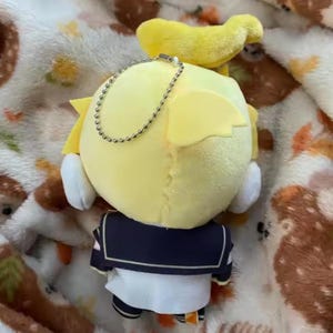May include: A yellow plush doll with a sailor uniform. The doll has yellow hair and a small yellow accessory on top. A silver chain is attached to the doll. The doll is on a patterned blanket.
