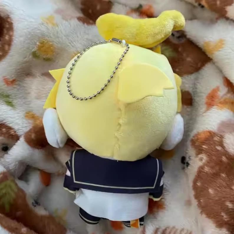 May include: A yellow plush doll with a sailor uniform. The doll has yellow hair and a small yellow accessory on top. A silver chain is attached to the doll. The doll is on a patterned blanket.