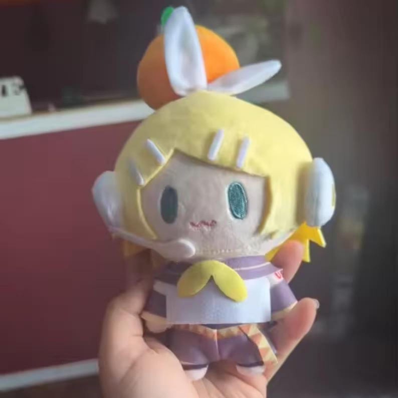 May include: A plush doll with yellow hair, a purple and white outfit, and a small orange hat with white bunny ears. The doll has large green eyes and a small embroidered mouth. The doll is held in a hand.