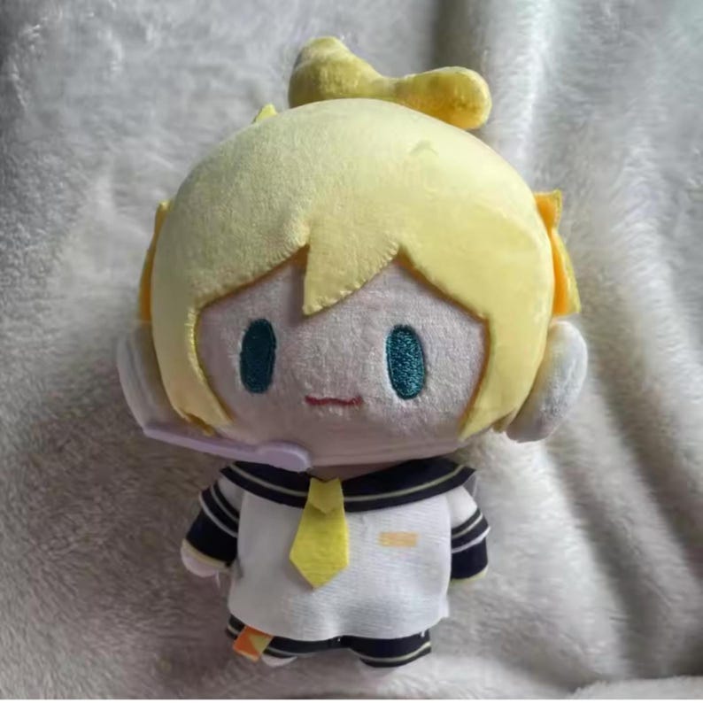 May include: A plush doll featuring a character with yellow hair, teal eyes, and a small red mouth. The doll wears a white sailor-style uniform with navy blue accents and a yellow tie. A headset is attached to the doll's head.