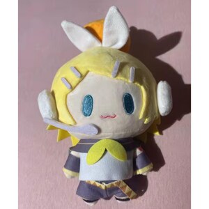 May include: A plush doll with a yellow head and hair, blue eyes, and a small red mouth. The doll has white ear-like features and is wearing a blue and yellow outfit. The doll is on a pink background.