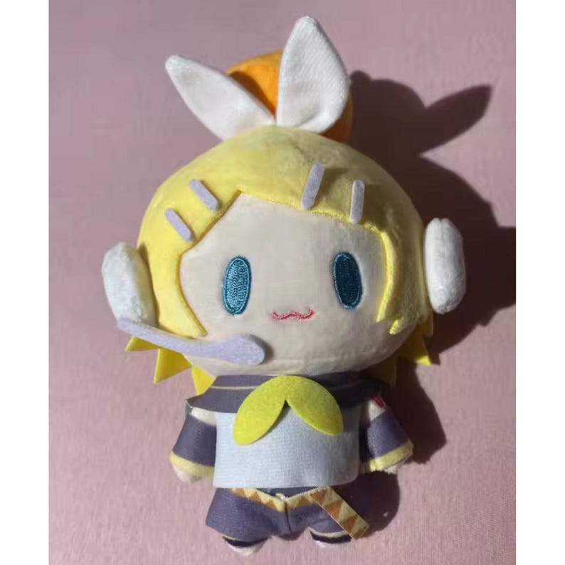 May include: A plush doll with a yellow head and hair, blue eyes, and a small red mouth. The doll has white ear-like features and is wearing a blue and yellow outfit. The doll is on a pink background.