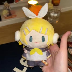 May include: A plush doll with yellow hair, blue eyes, and a small red mouth. The doll has a white and yellow outfit with a bow tie. The doll is holding a small wooden sword. The doll has white ear-like accessories and a white bow on top of its head.