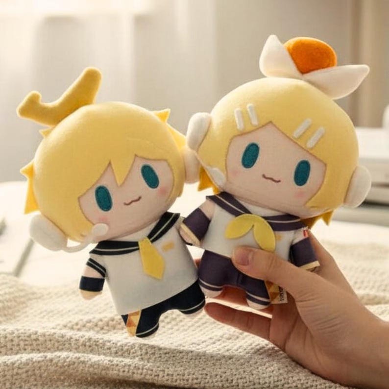 May include: Two plush dolls with yellow hair and sailor-style outfits. The dolls have light skin, blue eyes, and small smiles. One doll is held by a hand. The dolls have white and navy blue outfits with yellow accents.