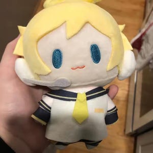 May include: A plush doll with yellow hair and a sailor-style outfit. The doll has embroidered blue eyes and a small, stitched orange mouth. The outfit is white with navy blue accents and a yellow tie. The doll is held in a person's hand.