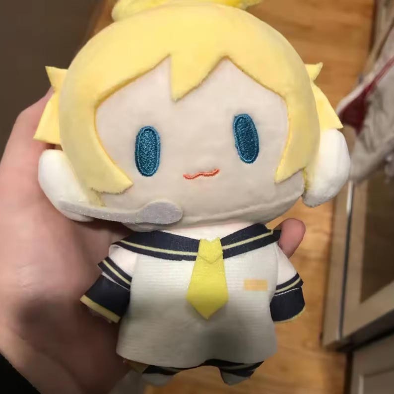 May include: A plush doll with yellow hair and a sailor-style outfit. The doll has embroidered blue eyes and a small, stitched orange mouth. The outfit is white with navy blue accents and a yellow tie. The doll is held in a person's hand.