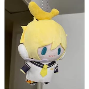 May include: A plush doll with yellow hair and a banana-shaped accessory. The doll wears a white sailor-style uniform with a yellow tie and navy blue accents. It has teal eyes and a small microphone.