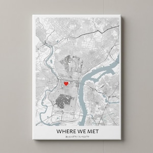 May include: A rectangular wall art print featuring a grayscale street map with a red heart icon marking a specific location. Below the map, the text reads WHERE WE MET followed by the coordinates 39.95258°N / 75.16524°W in black lettering.