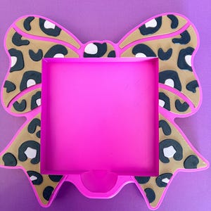 May include: A square-shaped frame with a bright pink border and a central cutout. The frame is decorated with a tan and black leopard print pattern shaped like a large bow, featuring raised black spots and white accents on the bow sections.