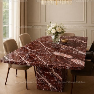 May include: A rectangular red marble dining table with white veining, supported by a matching pedestal base. The table is surrounded by four upholstered chairs. A vase of white flowers and a gold bowl sit on the table. A gold chandelier hangs above.