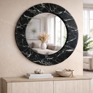 May include: A round black marble mirror with white veining, mounted on a beige wall. The mirror's frame has a polished finish. The mirror reflects a room with a beige sofa and a vase of dried flowers. The mirror is a decorative home decor item.