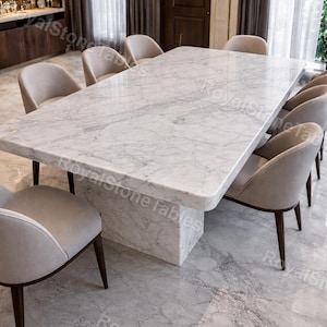 Puede incluir: Una gran mesa de comedor rectangular de mármol con un patrón veteado gris claro, rodeada de ocho sillas tapizadas. La mesa tiene una base de mármol macizo. La habitación tiene suelo de mármol. La mesa mide unos 240 cm.