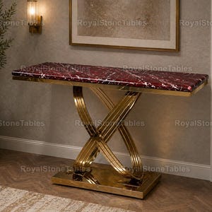 Puede incluir: Una consola rectangular con una tapa de mármol rojo y una base de metal dorado. La base presenta un diseño en forma de X. La mesa está colocada contra una pared de color neutro, con una planta y una obra de arte en el fondo.