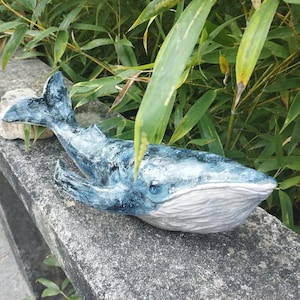May include: A handcrafted whale figurine featuring a textured surface with shades of deep blue, navy, and white. The sculpture displays a prominent dorsal fin, side flippers, and a wide tail fluke, with a smooth white underside and a visible eye.