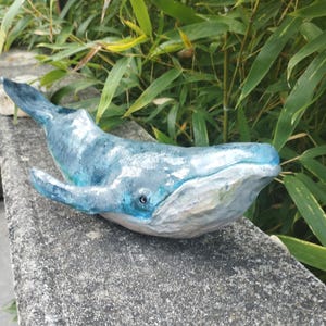 Peut inclure: Une figurine de baleine sculptée présentant une surface texturée avec des nuances de bleu, de blanc et de gris. La baleine possède une nageoire dorsale proéminente, un long corps et une large queue, avec un œil visible sur le côté de la tête.