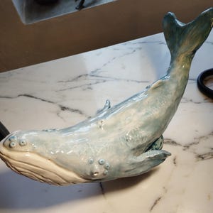 Puede incluir: Una escultura de ballena de cerámica con un acabado brillante en tonos azul claro y blanco. La ballena tiene una superficie texturizada con detalles circulares en relieve a lo largo del cuerpo, una aleta caudal y una parte inferior lisa color crema.
