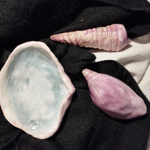 Peut inclure: Un ensemble de trois objets en céramique comprenant un bol ovale peu profond avec un intérieur bleu pâle et un bord rose, une pièce allongée en forme de coquillage strié dans des tons rose et blanc, et un objet lisse en forme de coquillage violet.