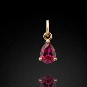 Puede incluir: Un colgante de oro con una gema roja oscura en forma de pera. La gema está engastada con tres pequeñas garras de oro. El colgante cuelga de un pequeño lazo de oro. El fondo es negro.