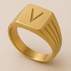 Könnte beinhalten: Goldfarbener Siegelring mit dem Buchstaben "V" auf der quadratischen Oberfläche. Der Ring hat eine gebürstete Metalloberfläche und drei vertikale Linien an der Seite. Ein klassisches Schmuckstück.