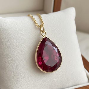 Könnte beinhalten: Eine goldene Halskette mit einem großen, tropfenförmigen Anhänger. Der Anhänger hat einen facettierten, tiefroten Edelstein, der in einer goldenen Lünette gefasst ist. Die Halskette wird auf einer weißen, gepolsterten Oberfläche in einer Holzbox präsentiert.