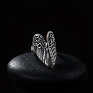 Op de afbeelding: Een zilverkleurige ring in de vorm van open vleugels. De ring heeft gedetailleerde veer- en schubpatronen tegen een donkere achtergrond. Een uniek sieraad.