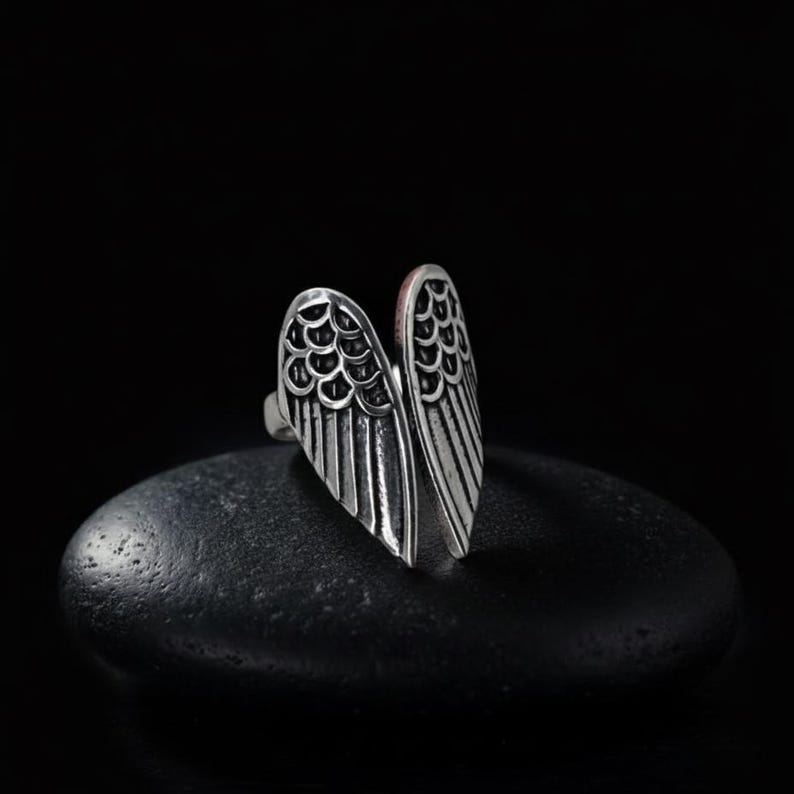 Op de afbeelding: Een zilverkleurige ring in de vorm van open vleugels. De ring heeft gedetailleerde veer- en schubpatronen tegen een donkere achtergrond. Een uniek sieraad.