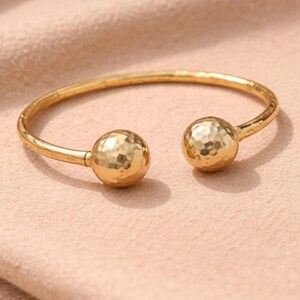 May include: A gold-colored open cuff bracelet. The bracelet features two textured, spherical end caps. The metal has a hammered appearance. The bracelet is displayed on a light pink surface.