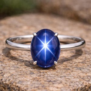 Könnte beinhalten: Ein silberner Ring mit einem großen, ovalen, tiefblauen Edelstein. Der Edelstein zeigt ein sternförmiges Lichtmuster. Der Ring liegt auf einer strukturierten, hellbraunen Oberfläche. Schmuck.